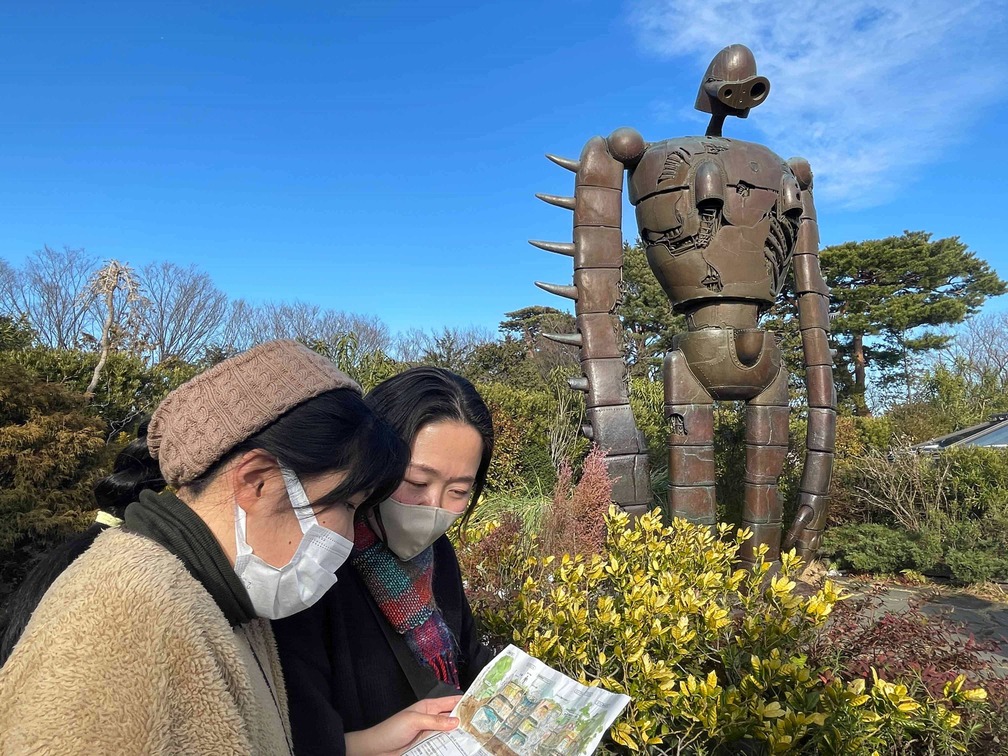 ジブリ美術館の屋上庭園のロボット兵の前で会場パンフレットを広げる関根さんと平澤さん。